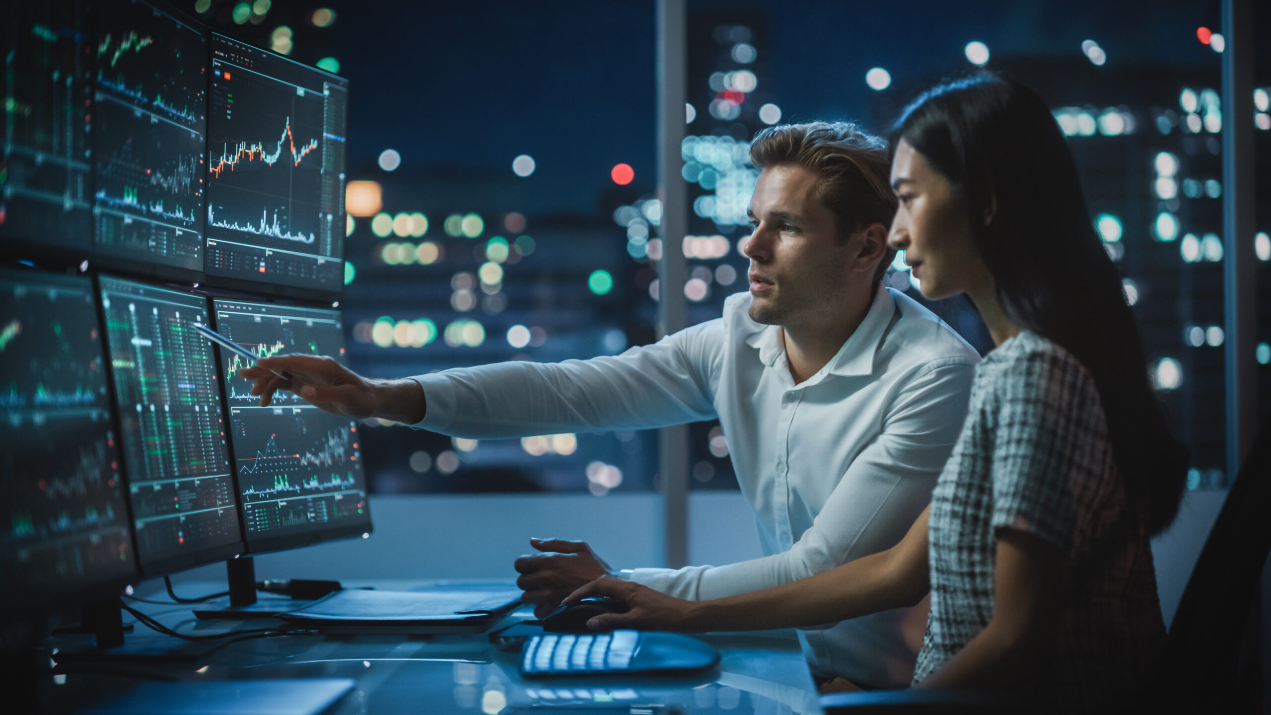 Financial Analyst Talking to Investment Banker in Late Evening at Work. Chatting About Real-Time Stock Chart Data on Desktop Computer. Businesspeople Have a Meeting in Broker Agency City Office.